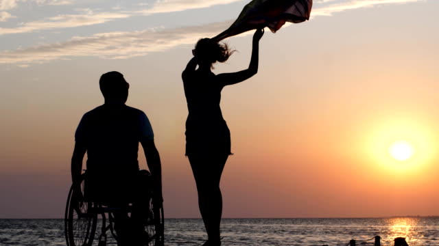 langbeinige-Frau-steht-auf-Steg-und-winken-ein-Tuch-vor-Kerl-verkrüppelt-in-Rollstuhl-auf-Grund-von-orange-Sonnenuntergang-über-Fluss