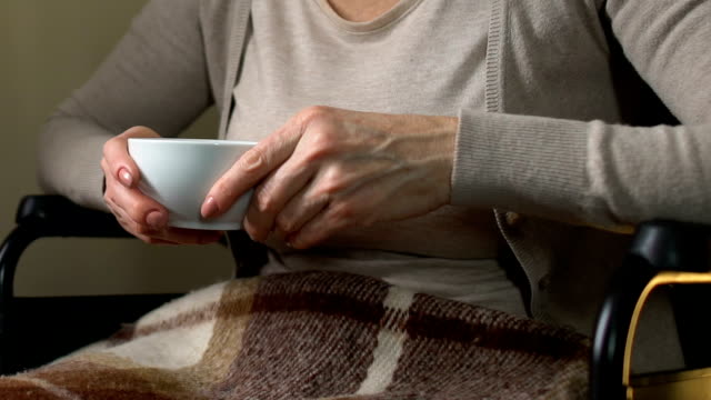 Mature-woman-sitting-near-window-in-wheelchair-drinking-hot-tea,-home-coziness