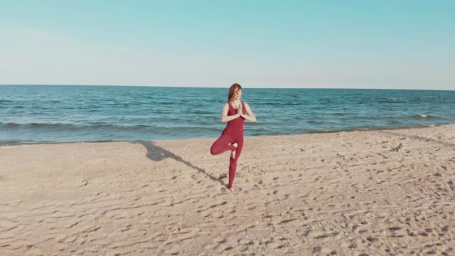 Drone-bewegt-sich-hinter-Frau-in-Sport-Wear-hält-Yoga-Asana-Position-am-Meer-oder-Ozean-Sandstrand.-Windige-sonniges-Wetter.-Luftaufnahme-des-friedlichen-Gesundheit-Mädchen-Praxis-durchführen