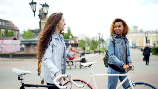 Cheerful-young-women-are-talking-while-walking-with-bikes-along-beautiful-street-with-fountain-and-beautiful-buildings-and-people-moving-in-background.