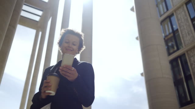 Tracking-left-shot-of-middle-adult-female-manager-with-short-hair-standing-outdoors-on-windy-day,-holding-coffee-cup-and-texting-on-her-mobile-phone-with-smile