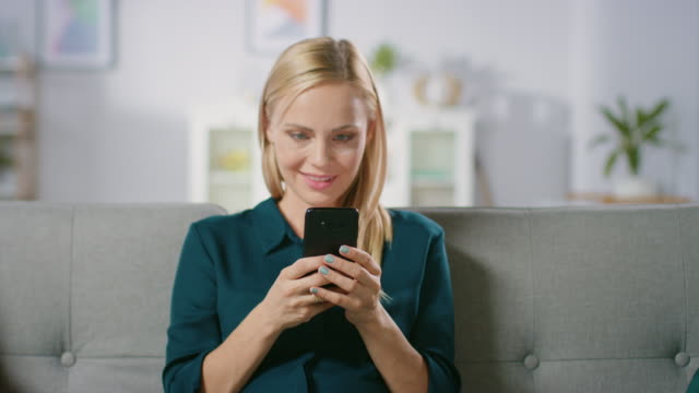 Hermosa-rubia-mujer-utiliza-teléfono-móvil-mientras-está-sentado-en-un-sofá-en-casa.-Sonriendo-feliz-mujer-utiliza-el-Smartphone-para-navegar-a-través-de-Internet,-redes-sociales-y-viendo-Videos.