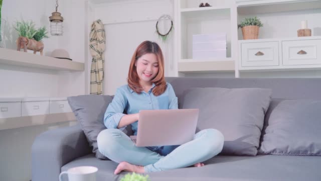 Porträt-der-schönen-attraktiven-jungen-lächelnden-Asiatin-mit-Computer-oder-Laptop-auf-dem-Sofa-liegend-beim-Entspannen-im-Wohnzimmer-zu-Hause.-Genießen-Zeit-Lifestyle-Frauen-bei-Wohnkonzept.