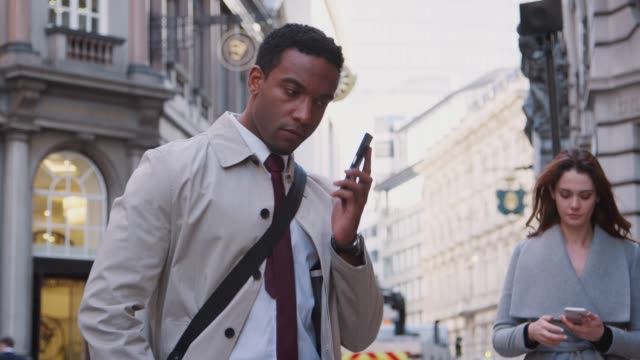 Young-black-businessman-standing-on-street-talking-on-smartphone,-white-millennial-woman-walking-past-also-using-phone,-close-up,-low-angle