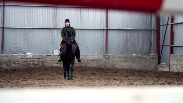 in-special-hangar,-a-young-disabled-man-learns-to-ride-a-black,-thoroughbred-horse,-hippotherapy.-man-has-an-artificial-limb-instead-of-his-right-leg.-concept-of-rehabilitation-of-disabled-with-animals