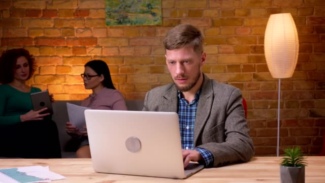 Closeup-shoot-of-adult-businessman-using-the-laptop-looking-at-camera-and-smiling-indoors-in-the-office.-Female-employee-discussing-data-with-a-colleague-on-the-background
