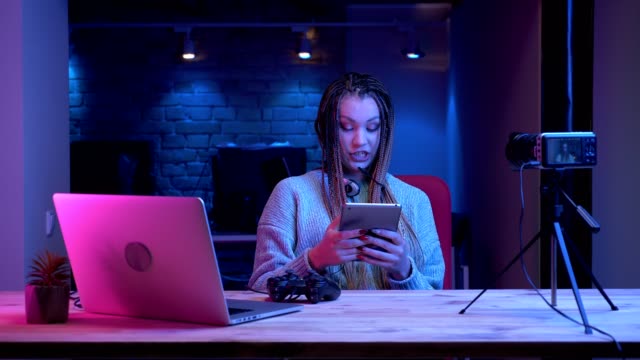 Closeup-shoot-of-young-attractive-female-blogger-with-dreadlocks-in-headphones-streaming-live-talking-about-video-games-using-the-tablet-with-the-neon-background-indoors