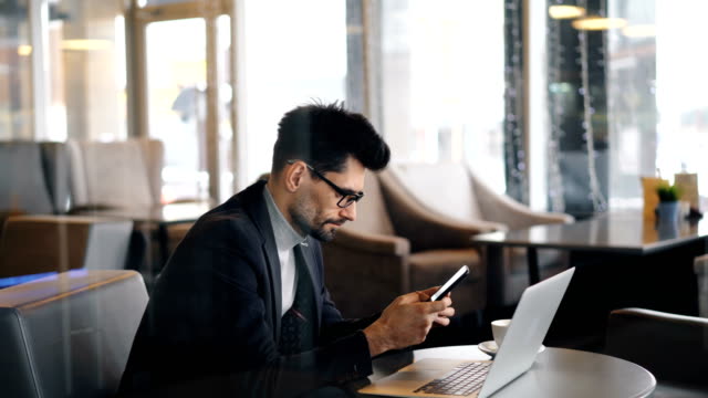 Confident-bearded-man-in-glasses-using-smartphone-during-coffee-break-in-cafe