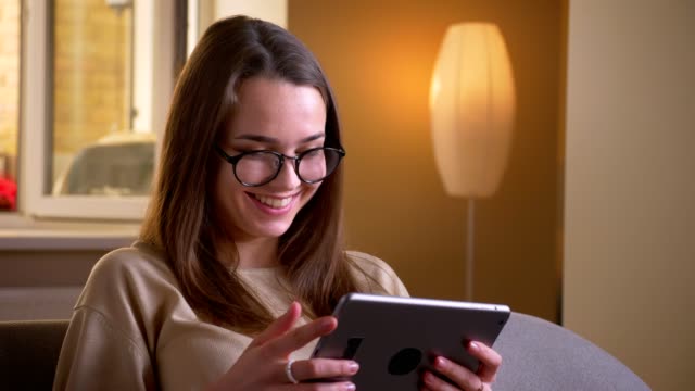 Closeup-portrait-of-young-attractive-caucasian-female-in-glasses-using-the-tablet-smiling-happily-sitting-on-the-couch-indoors-in-apartment