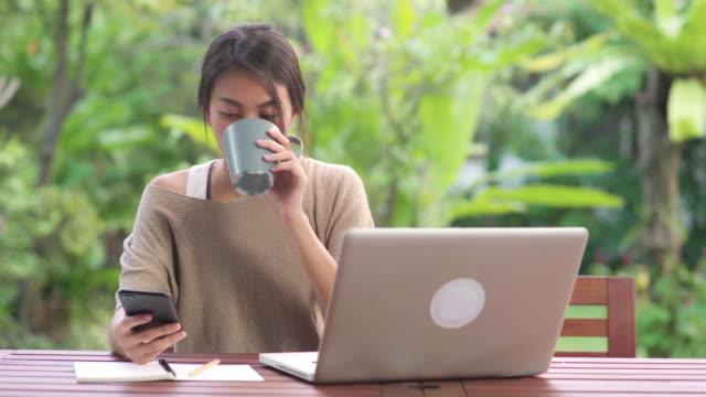 Freelance-Asian-woman-working-at-home,-business-female-working-on-laptop-and-using-mobile-phone-drinking-coffee-sitting-on-table-in-the-garden-in-morning.-Lifestyle-women-working-at-home-concept.