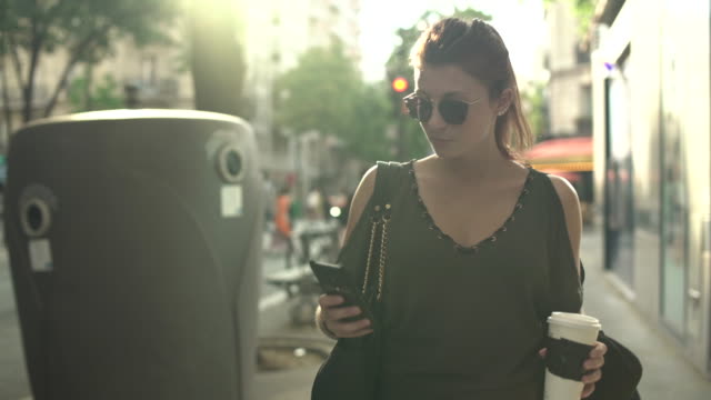 Attraktive-lächelnde-kaukasische-Frau-mit-Sonnenbrille,-Sommersprossen,-Piercings-und-roten-Haaren-schreiben-SMS-an-Freund-auf-ihrem-Smartphone-und-Kaffee-trinken,-die-durch-die-Straße-in-Paris-gehen.-4K.