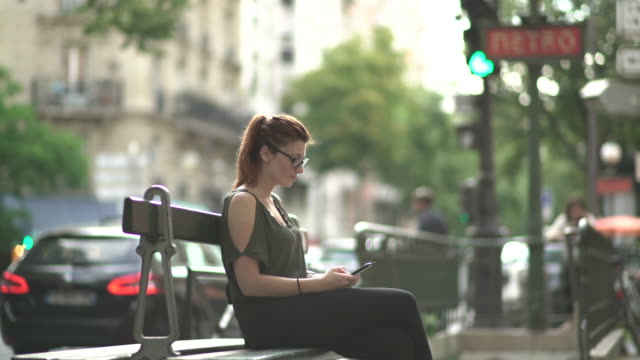 Attraktive-kaukasisch-nachdenkliche-Frau-mit-Brille,-Sommersprossen,-Piercings-und-roten-Haaren-warten-und-beobachten-an-ihrem-Smartphone-sitzen-auf-der-Bank,-während-sonnigen-Sommer-in-Paris.-4K-UHD.
