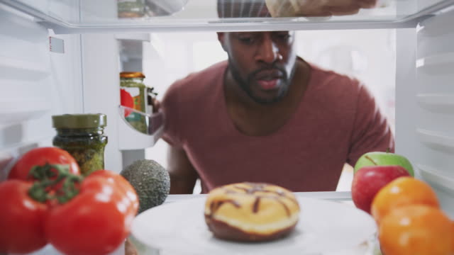Blick-aus-dem-Inneren-des-Kühlschranks-als-Mann-öffnet-Tür-und-greift-nach-ungesunden-Donut