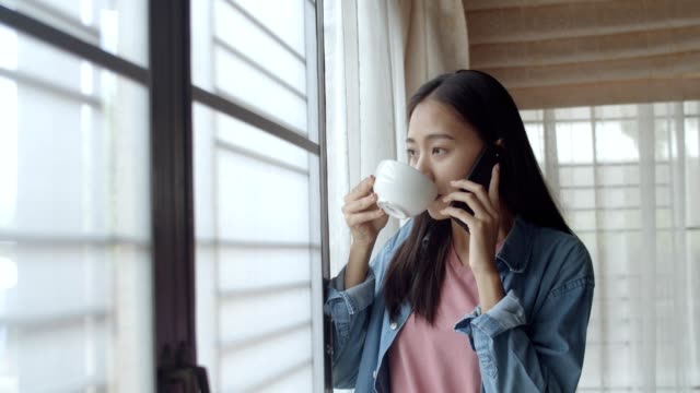 Atractivo-retrato-sonriente-joven-asiática-mujer-bebe-café-y-hablando-por-teléfono-con-amigos-de-pie-al-lado-de-la-ventana-en-la-oficina-en-casa.