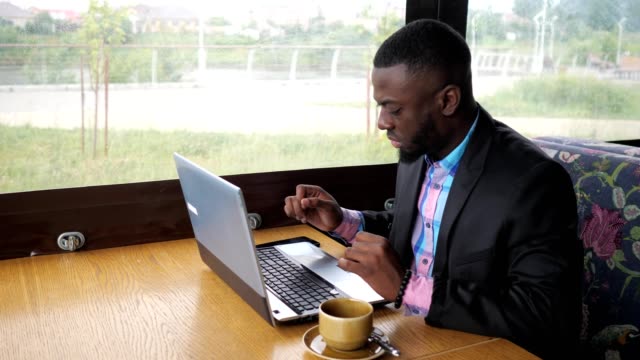 Black-businessman-is-working-typing-a-message-on-laptop-sitting-in-summer-cafe.