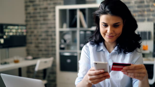 Chica-alegre-comprando-en-línea-pagando-con-tarjeta-de-crédito-usando-el-teléfono-inteligente-en-la-oficina