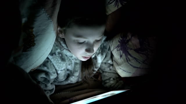 Cute-little-girl-at-night-in-the-dark-playing-on-the-tablet-under-the-blanket-on-the-bed.-Concept-video.-Close-up.-Raw-video.-4K.