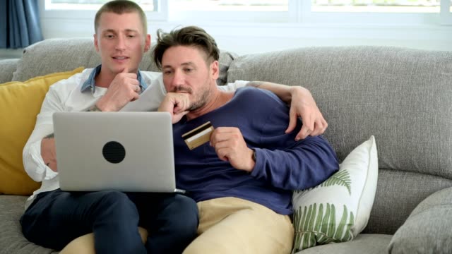 Gay-couple-relaxing-on-couch-using-laptop-computer.-Confuse-mood.