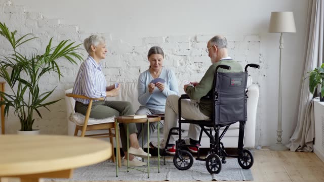 Group-of-three-elderly-people,-two-senior-women-and-disabled-old-man-in-wheelchair,-playing-cards-in-common-room-of-nursing-home,-wide-shot