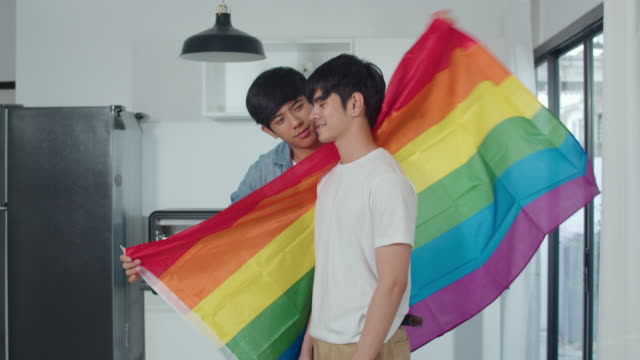 Pareja-gay-asiática-de-pie-y-habitación-de-abrazo-en-casa.-Jóvenes-hombres-LGBTQ+-guapos-besando-feliz-relax-descanso-juntos-pasar-tiempo-romántico-en-la-cocina-moderna-con-bandera-arco-iris-en-casa-en-el-concepto-de-la-mañana.