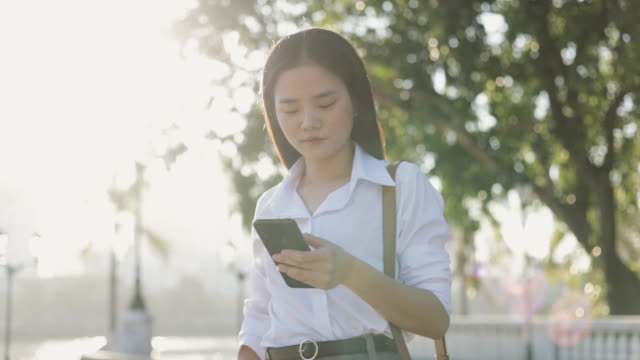 Schöne-asiatische-Geschäftsfrau-in-einem-weißen-Hemd-verwendet-ein-Smartphone-SMS-Teilen-von-Nachrichten-auf-Social-Media,-während-außerhalb-des-Morgens-in-einem-öffentlichen-Park.