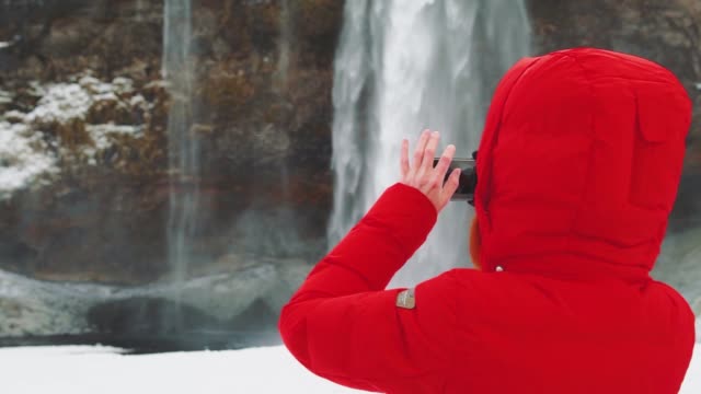 schöne-junge-Frau-in-der-Nähe-eines-Wasserfalls-in-Island,-macht-Fotos-am-Telefon