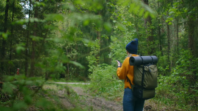 Slow-motion:-Young-woman-hiker-using-her-smart-phone-while-enjoying-her-weekend-in-forest.-Traveling-woman-with-backpack-walking-on-path-the-forest-looking-at-the-telephone-and-map-in-green-wood