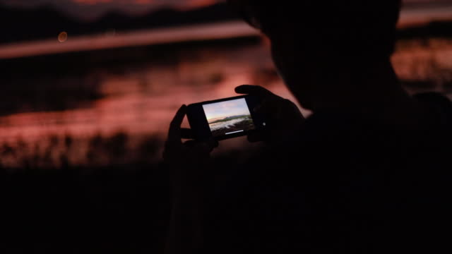 Silhouette-von-Nahaufnahme-asiatischen-Mann-mit-Smartphone-überprüfen-ein-Foto-nach-dem-Fotografieren-am-Meer-Strand-schönen-Sommer-Sonnenuntergang,-goldene-Stunde-Licht.