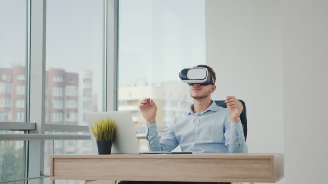 A-young-man-sitting-at-a-desk-in-the-office-uses-augmented-reality-glasses-to-work-on-business-projects-in-various-fields.