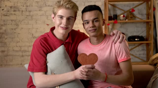 Multiethnic-happy-gay-couple-sitting-on-the-couch-and-holding-a-heart-sign-in-hands.-Homeliness,-LGBT-loverHappy-s,-happy-gay-family-concept.-60-fps