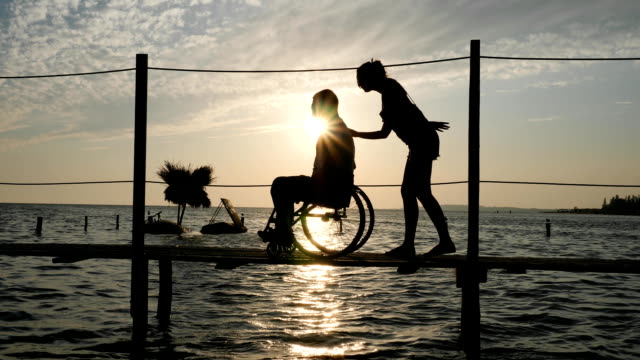 shapely-female-with-guy-invalid-on-wheel-chair-going-on-jetty-against-heaven-in-sundown
