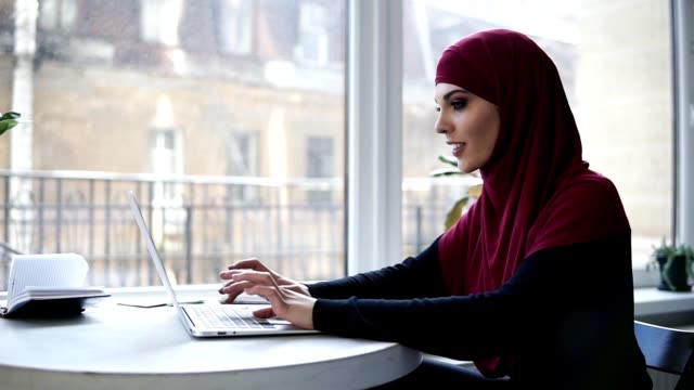 Junges-attraktives-muslimische-Mädchen-mit-Hijab-ihren-Kopf-zu-bedecken-ist-etwas-auf-ihrem-Laptop-die-Eingabe-während-der-Sitzung-in-einige-angeblich-Coworking-Raum-mit-Fenstern-auf-dem-Hintergrund