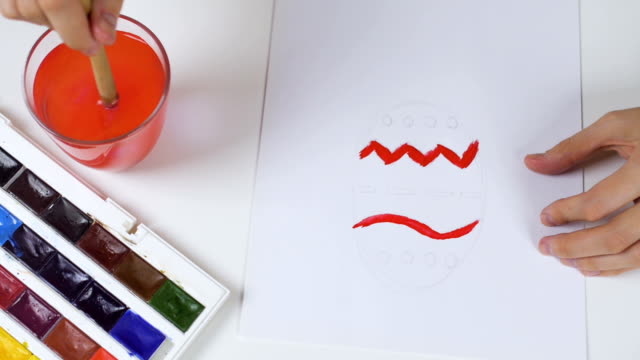 Unrecognizable-woman-paints-a-sketch-with-bright-colors-on-paper-for-an-Easter-card.-On-the-table-is-a-palette-with-paints-and-a-glass-of-water.
