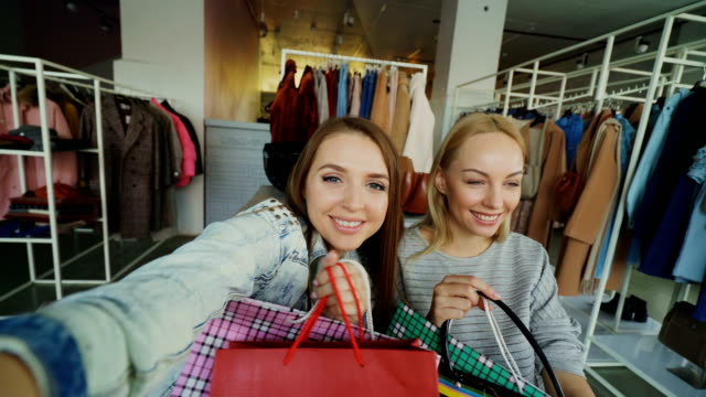Point-of-view-shot-of-attractive-young-ladies-making-selfie-with-paper-bags-in-women's-clothes-boutique.-Girls-are-posing,-chatting-and-laughing-happily