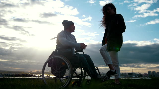 Hombre-discapacitado-en-silla-de-ruedas-con-mujer-hablando-en-la-puesta-de-sol-nublado