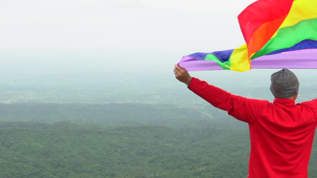 man-raise-rainbow-colour-LGBTI-flag-waving-in-hard-wind-on-mountain-top-viewpoint