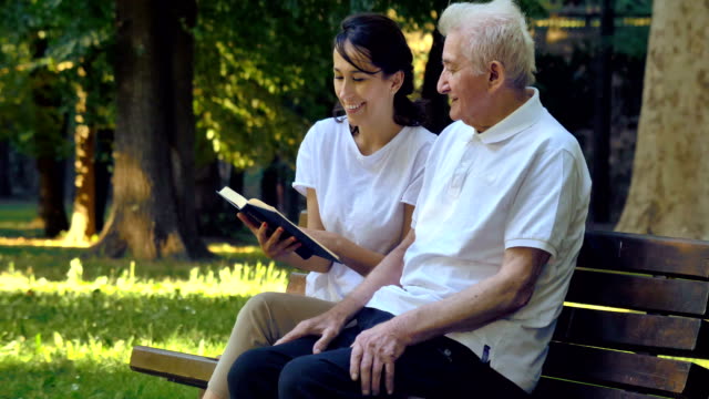 Ein-Kindermädchen,-eine-Krankenschwester,-Fürsorge-für-ältere-Menschen,-ein-Mädchen-(Frau)-und-Großvater-auf-ein-Taschenbuch-sitzen-und-ein-Buch,-im-Park-lesen.