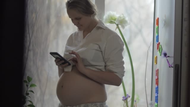 Lächelnde-schwangere-Frau-mit-Handy-zu-Hause