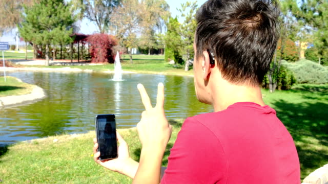 Young-deaf-boy-communicating-with-sign-language--at-smart-phone-in-park