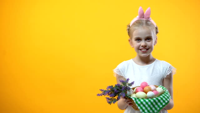 Lächelnde-Kind-zeigt-Körbchen-mit-bunten-Eiern-gelben-Hintergrund,-Ostern