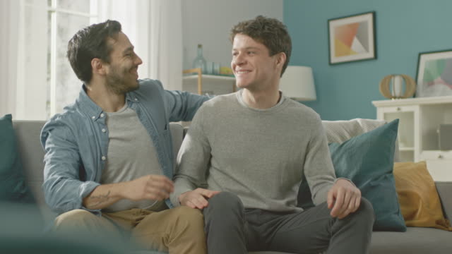 Cute-Attractive-Male-Queer-Couple-Sit-Together-on-a-Sofa-at-Home.-Gay-Boyfriend-Puts-His-Hand-on-Partner's.-They-Hug.-It's-a-Happy-Relationship-and-They-Smile.-Room-Has-Modern-Interior.