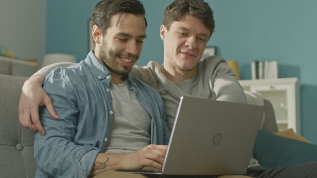Sweet-Male-Gay-Couple-Spend-Time-at-Home.-They-Sit-on-a-Sofa-and-Use-the-Laptop.-They-Browse-Online.-Partner's-Hand-is-Around-His-Lover.-Room-Has-Modern-Interior.