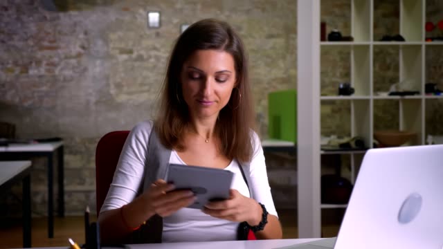 Attractive-female-office-worker-taps-twice-on-the-screen-of-tablet-smiles-and-shows-the-image-to-a-camera