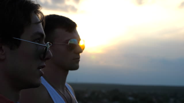 Profile-of-handsome-gay-boys-in-sunglasses-standing-on-the-edge-of-rooftop-and-talking.-Young-male-couple-relaxing-on-roof-of-high-rise-building-and-enjoying-beautiful-cityscape.-Close-up-Slow-motion