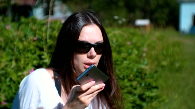 Beautiful-brunette-woman-records-a-voice-message-on-her-mobile-phone-while-sitting-in-the-park-on-a-Sunny-day.