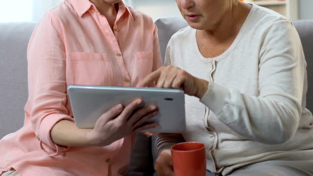 Adult-daughter-teaching-old-mom-to-use-tablet,-showing-video-call-application