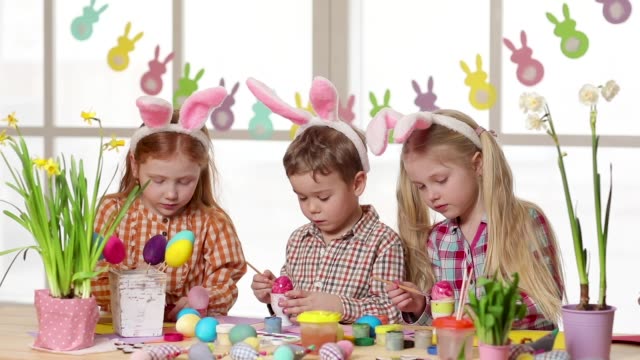 Niños-felices-usando-orejas-de-conejo-pintando-huevos-el-día-de-Pascua.-Niñas-pequeñas-preparándose-para-la-Pascua.