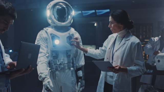 Diverse-Team-of-Aerospace-Engineers-Design-New-Space-Suit-Adapted-for-Galaxy-Exploration-and-Travel.-Group-of-Scientists-Wearing-White-Coats-have-Discussion,-Use-Computers.-Constructing-Astronaut-Suit