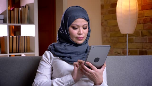 Tiro-de-primer-plano-de-la-hembra-musulmana-adulta-en-hijab-usando-la-tableta-mientras-está-sentado-en-el-sofá-en-el-interior-de-su-casa
