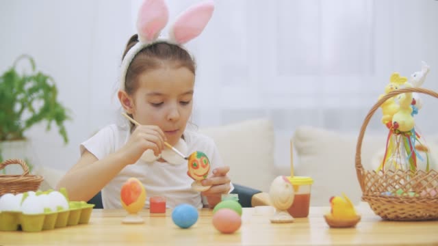 Little-playful-girl,-wearing-bunny-ears-on-her-head-chose-an-orange-colour-to-paint-an-egg.-Girl-is-chewing-and-outting-a-yellow-spot-on-her-nose.